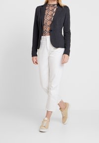 Blazer ajustado gris oscuro sobre una blusa beige con estampado de serpiente, combinado con pantalones blancos cortos y zapatillas beige. Fondo liso.
