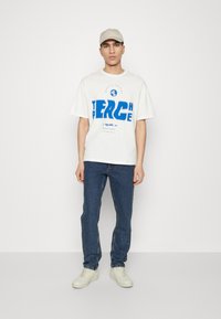 Camiseta gráfica blanca con texto azul, corte relajado, combinada con jeans azules y zapatillas blancas. Modelo lleva una gorra beige, fondo neutro.