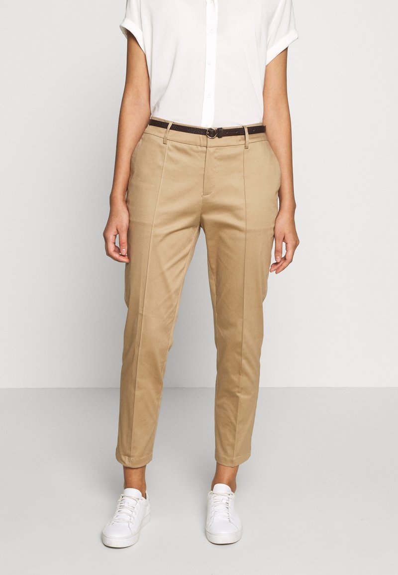 Femme portant un pantalon beige sur mesure avec des coutures à l'avant, une ceinture foncée, une chemise blanche à manches courtes boutonnée, et des baskets blanches, debout devant un fond blanc.