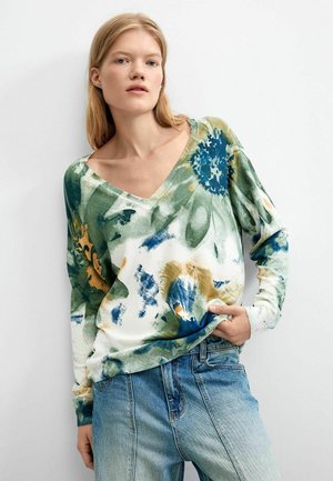 Joven con cabello rubio que lleva un suéter con cuello en V y estampado floral en verde, azul y amarillo, y vaqueros azul claro.