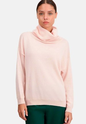 Pull à col roulé rose en tricot doux, avec des manches longues et des accents côtelés, associé à un pantalon vert foncé.