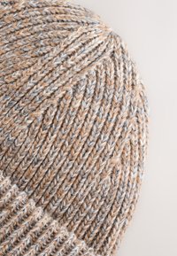 Knitted beanie in tan and gray, featuring a ribbed texture and a rounded shape. Contains varied yarn thickness for visual interest.