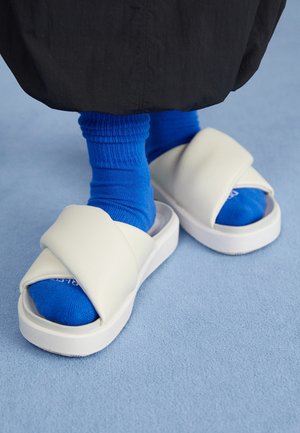 Stopy w jasnoniebieskich skarpetkach i białych, wyściełanych sandałach typu slide na jasnoniebieskiej podłodze, z czarnym materiałem widocznym powyżej kostek.