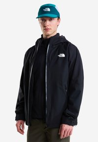 Νέος άνδρας που φοράει μαύρο μπουφάν The North Face και τιρκουάζ καπέλο The North Face, στέκεται μπροστά σε απλό λευκό φόντο.