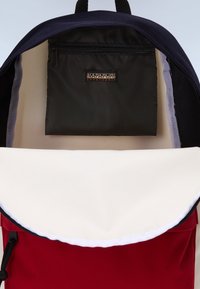 L'intérieur d'un sac à dos en tissu rouge et blanc présente une poche zippée foncée et un dessus bleu marine. Des coutures contrastantes sont visibles autour de l'ouverture.