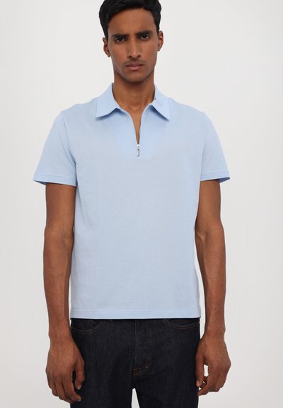 Απαλό μπλε πόλο μπ�λουζάκι με κοντό φερμουάρ, επίπεδο γιακά και κοντά μανίκια. Μαλακή υφή, εφαρμοστή γραμμή, συνδυασμένο με σκούρα τζιν.