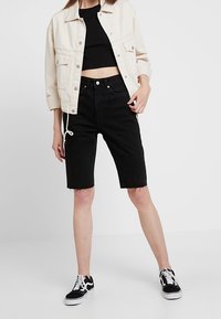 Zwarte denim shorts met een rafelige rand, gecombineerd met een zwart crop top en een crèmewitte oversized jas. Zwart-witte sneakers maken de outfit compleet.