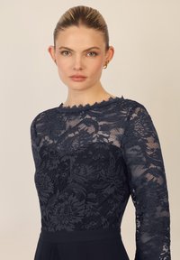 Vestido de encaje azul marino con mangas largas, patrones florales, escote con forma de ola y un corsé ajustado que se transforma en una falda sólida.