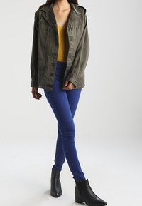 Veste légère vert olive avec un grand col, des poches avant et des détails zippés, associée à un haut jaune et un jean skinny bleu. Bottines noires.