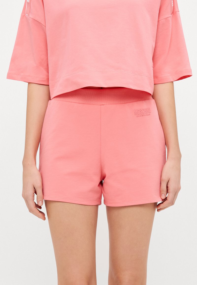 Pink cotton shorts with a ribbed waistband feature a logo embossed on the front. The fabric appears soft and casual in design.