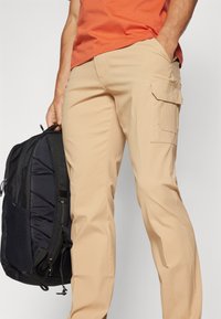 Beige cargo bukser med sidelommer, lavet af letvægtsstof. En sort rygsæk i venstre hånd, der har et struktureret design.