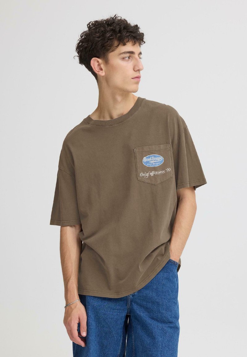 T-shirt en coton marron à manches courtes, avec une poche poitrine et un détail de logo brodé. Porté avec un jean en denim bleu.