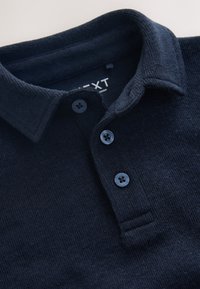 Polo en maille marine avec col côtelé, fermeture à trois boutons et tissu texturé. Étiquette de la marque visible à l'intérieur du col.