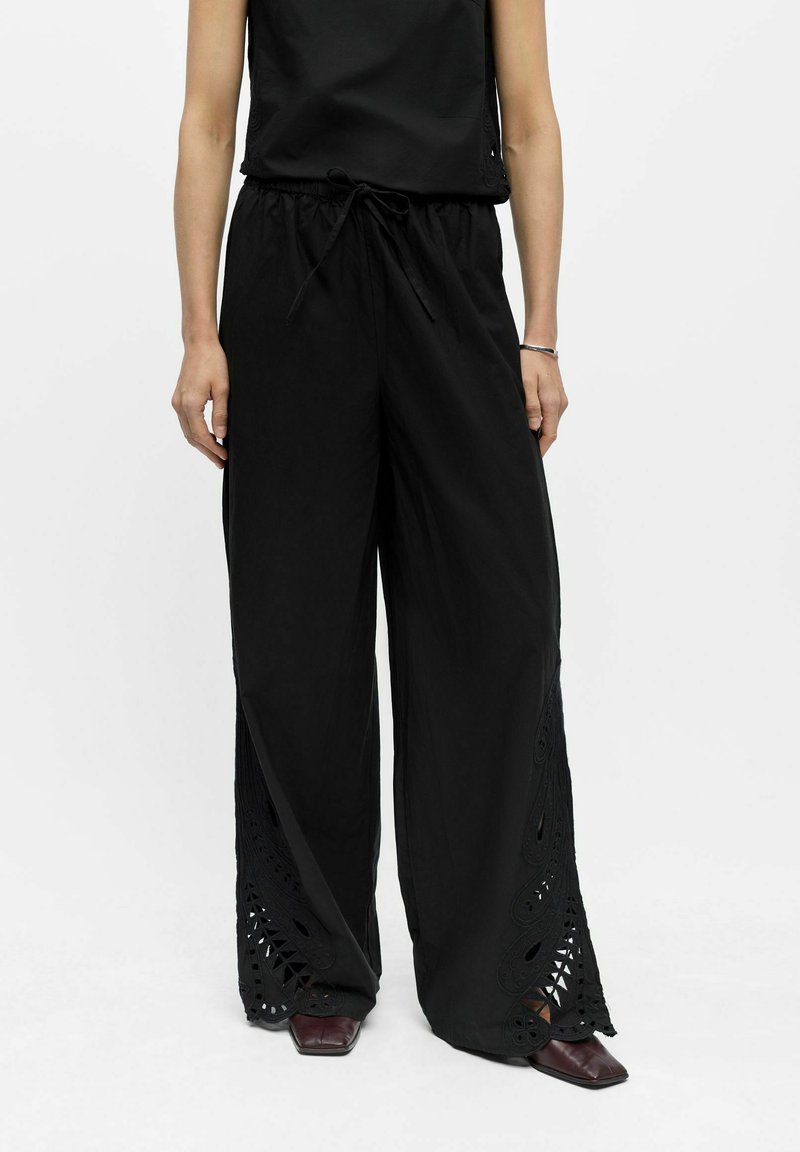 Femme portant un pantalon noir à jambes larges avec une taille à cordon et des découpes en dentelle près de l'ourlet, assorti à des chaussures foncées à bout carré.