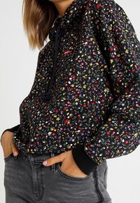Sudadera negra con estampado floral, capucha con cordón, puños y dobladillo elásticos. El material parece suave con una textura ligera.