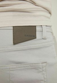 Pantalon gris clair avec un patch en cuir texturé portant l'inscription "CHASIN'" à l'arrière. Comprend des détails cousus et des poches arrière.