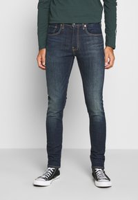 Hombre vistiendo jeans oscuros ajustados y zapatillas altas negras, de pie frente a un fondo sencillo.