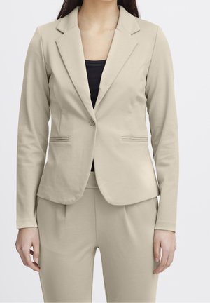 Frau trägt einen beige maßgeschneiderten Blazer und passende Hose über einem schwarzen Top, steht vor einem einfarbigen hellen Hintergrund.