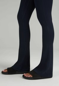 Czarne dzwony legginsy o gładkiej fakturze i kontrastowych szwach, zestawione z czarnymi klapkami z logo na pasku.