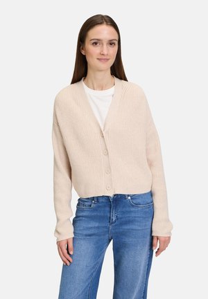 Cardigan beige à côtes avec un col en V, manches longues et fermeture à cinq boutons, porté sur une chemise blanche et associé à un jean bleu.