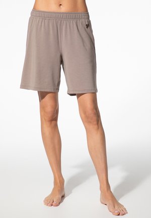 Person, die locker sitzende, taupefarbene Shorts mit elastischem Bund trägt, barfuß auf hellem Hintergrund steht, Beine sind von der Taille abwärts sichtbar.