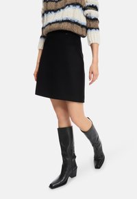 Schwarze knielange Rock mit tailliertem Bund und glatter Textur, kombiniert mit hohen schwarzen Lederstiefeln mit eckiger Spitze und Blockabsatz.