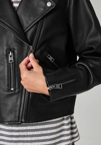 Crna kožna motoristička jakna s više zip zatvarača, oštar ovratnik i uski kroj. Ispod se vidi siva prugasta košulja.