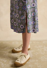 Grün gemustertes Kleid mit Blumen, seitlichem Schlitz und beige strukturierten Sneakers, die eine braune Gummisohle und Schnürsenkel haben.