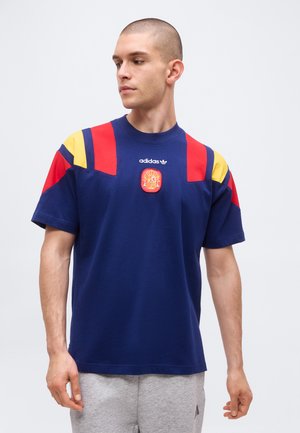 Homme portant un t-shirt bleu marine Adidas avec des bandes rouges et jaunes aux épaules et un écusson aux armoiries de l’Espagne, regardant vers sa gauche.