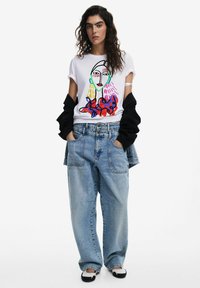 Weißes grafisches T-Shirt mit einem bunten abstrakten Gesichtdesign, kombiniert mit übergroßen hellblauen Jeans und schwarzen, gestuften Sweatshirtärmeln.