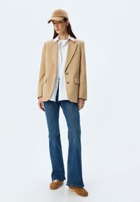 Frau mit einer beigen Mütze, beigem Blazer über einem weißen Hemd, blauen Jeans und braunen Sneakern, die vor einem schlichten weißen Hintergrund steht.