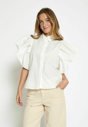 Camicia bianca a maniche corte con maniche voluminose arricciate, chiusura con bottoni e colletto classico. Indossata con pantaloni beige a vestibilità ampia.