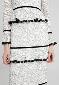 Robe en dentelle blanche avec bordure noire et détails pompons sur les manches, la taille et la jupe, portée par une personne avec les mains détendues sur les côtés.