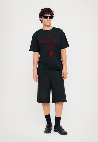 T-shirt en coton noir avec du texte rouge et un graphique de rose, associé à des shorts noirs et des mocassins noirs. Le mannequin porte des lunettes de soleil noires.