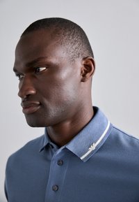 Blå polo skjorte med teksturert overflate, to hvite striper på kragen og en sølvfarget logoaccent. Knapper er synlige på knappestykket.