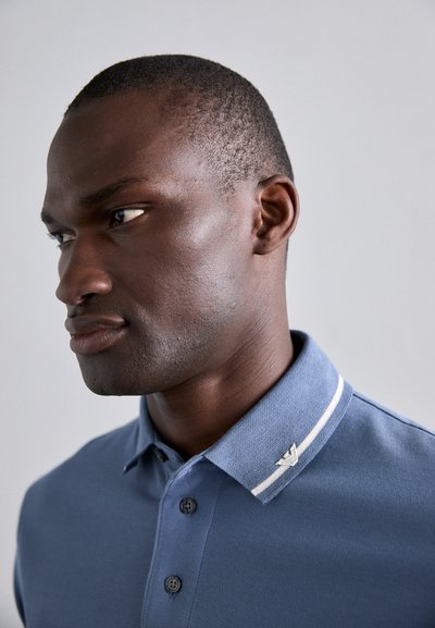 Tricou polo albastru cu un finisaj texturat, două dungi albe la guler și un accent de logo argintiu. Butoanele sunt vizibile pe brâul frontal.