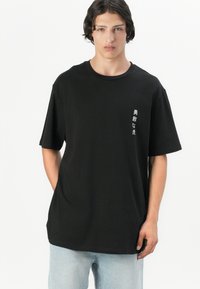 Schwarzes Baumwoll-T-Shirt mit kurzen Ärmeln, Rundhalsausschnitt und weißem Drucktext auf der linken Brust. Lockere Passform. Hellblaue Jeans.