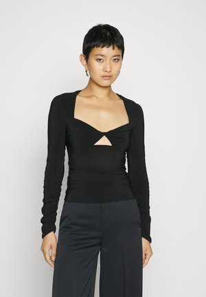 Νεαρός με κοντά μαύρα μαλλιά, φορώντας ένα μαύρο μακρυμάνικο μπλουζάκι με άνοιγμα στο στήθος και σκούρο παντελόνι σε φαρδιά γραμμή, μπροστά σε απλό φόντο.