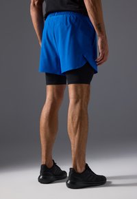 Zili zili sporta šorti ar šķeltu dizainu, saskaņoti ar melniem kompresijas šortiem. Modelis valkā melnas sporta kurpes.