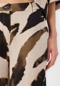 Pantalons beiges clairs avec un motif abstrait audacieux en noir et marron, dotés d'une fermeture à bouton à l'avant, de poches latérales et d'un tissu texturé.
