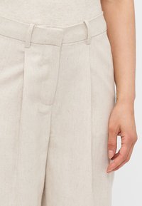 Pantalons en lin beige à taille haute, dotés de plis marqués, de passants de ceinture et d'une finition texturée. Une main repose sur le tissu.