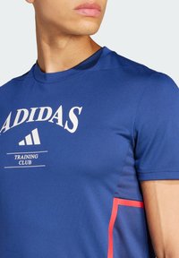 Blå träningströja med vit logotyp "ADIDAS TRAINING CLUB", som har en slät yta och kontrasterande röda detaljer på sidan.
