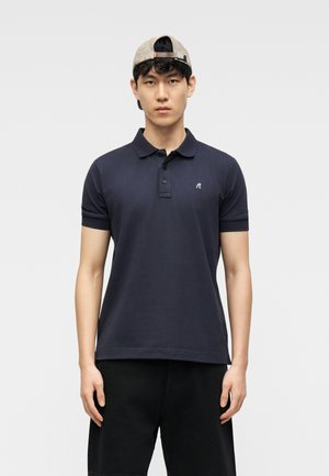 Mladý muž v tmavomodrom polokošeli a čiernych šortkách s béžovou šiltovkou nasadenou dozadu, stojaci na jednoduchom svetlom pozadí.