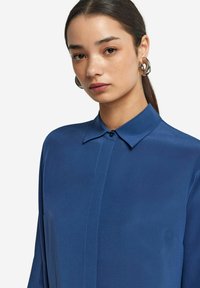 Blaue Satinbluse mit klassischem Kragen, Knopfleiste und langen Ärmeln. Glatte Textur mit dezentem Glanz.