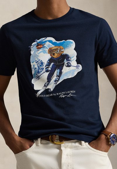 Navy cotton t-shirt featuring a graphic of a skiing bear, wearing goggles, against a blue and white snowy background. Text at the bottom identifies the brand.