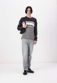Gray ribbed sweater with white and black stripes, paired with light gray jeans and black sneakers. Model holds a small black bag.