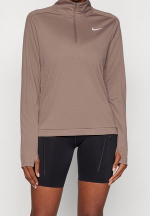 Bluza Nike z długim rękawem w kolorze taupe, z krótkim zamkiem i białym logo, zestawiona z czarnymi dopasowanymi spodenki sportowymi z kieszeniami na zamek.