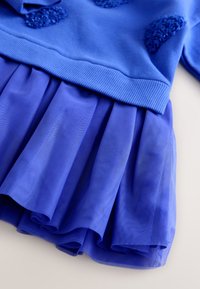 Kék pulóver texturált részletekkel és réteges szoknya áttetsző anyagból, pliszírozott mintával, amely a szegélyen lebeg.