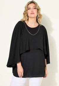 Haut noir à couches avec des manches chauve-souris, présentant un accent de chaîne délicat. Tissu lisse avec un design fluide, associé à un pantalon blanc.
