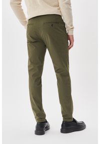 Olive green chinos with a slim fit, featuring two back pockets with black buttons. Smooth fabric and tapered legs. Black shoes worn.